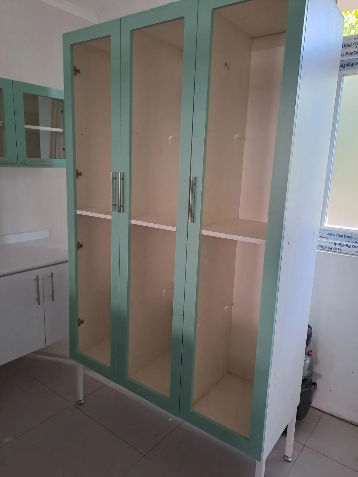 Mueble alto de dos cuerpos con marcos verde agua y puertas de vidrio, junto a un módulo bajo blanco