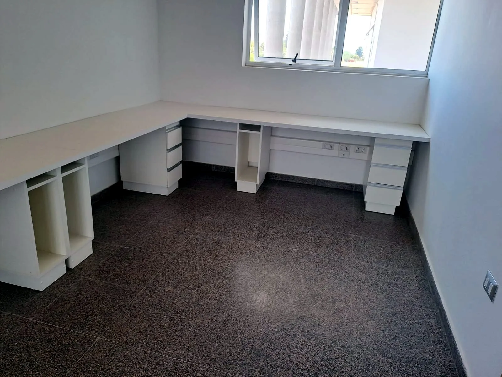 Oficina con escritorio blanco en L bajo la ventana, cajoneras y porta CPU