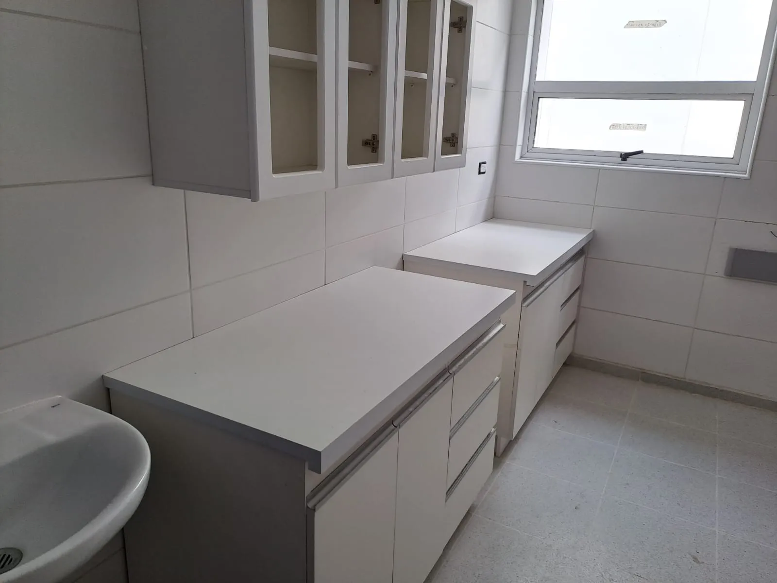 Muebles bajos blancos con cajones y cubierta blanca en L junto a un lavamanos, con aéreos de puertas de vidrio